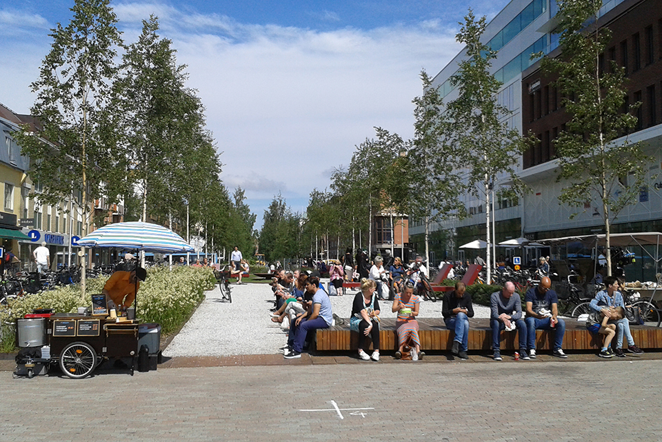 folk sitter i solen, träd, hus, korvförsäljning