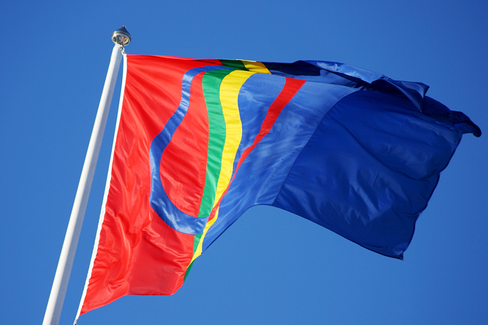 Samisk flagga vajar på flaggstång mot blå himmel.