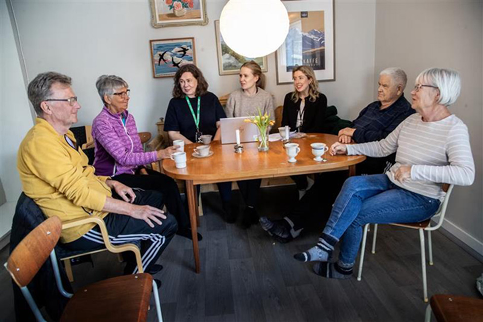 Personer sitter runt ett bord med tavlor i bakgrunden.