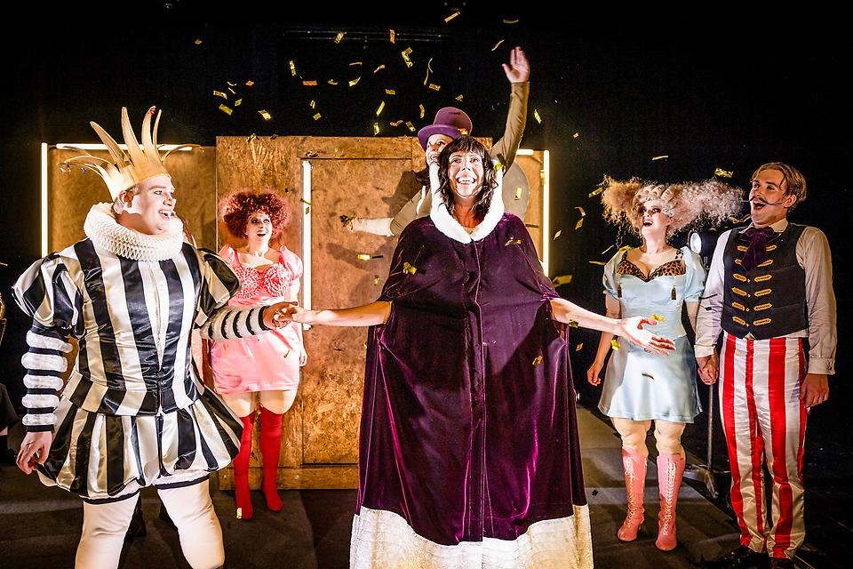 Personer i färgglada kläder i feststämning med konfetti i luften. En kvinna i vinröd kåpa i mitten. En man med krona på huvudet till vänster.