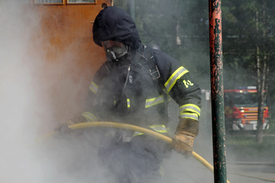 En brandman med rökdykarutrustning håller i en slang riktad in genom en dörröppning som det kommer rök ifrån. I bakgrunden syns en brandbil stå parkerad