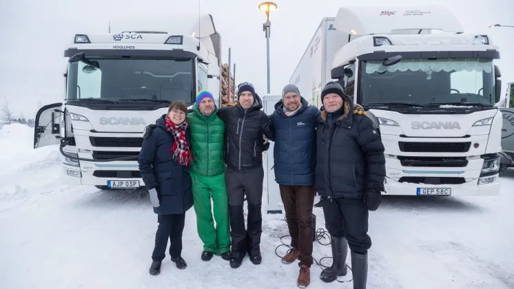 Fem personer står tillsammans framför två lastbilar om vintern.