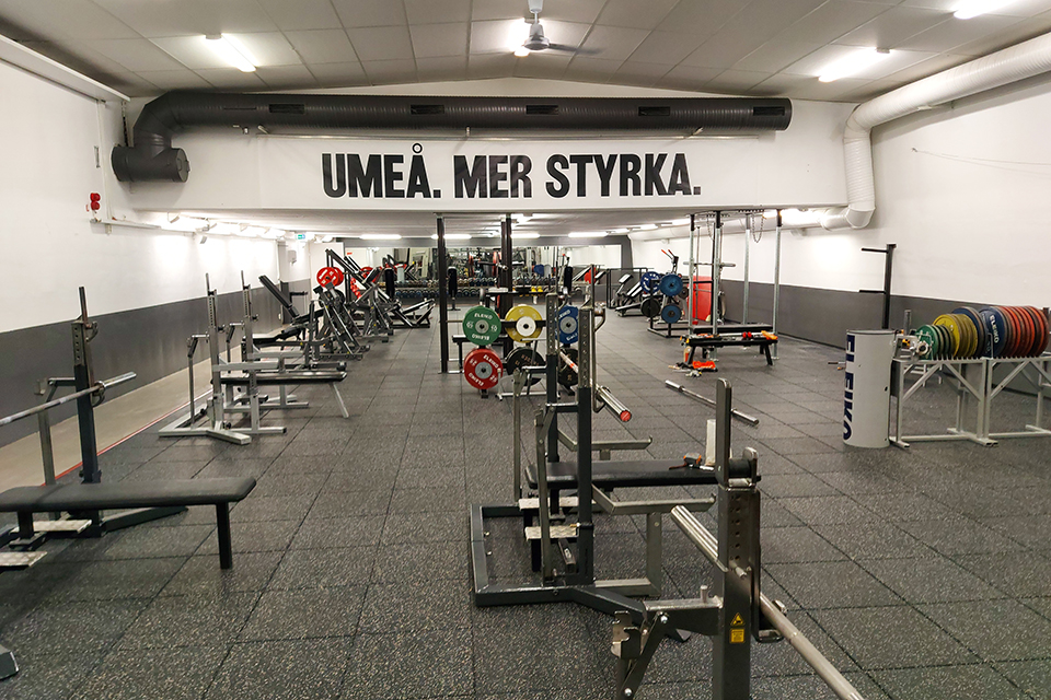 En banderoll med budskapet Umeå mer styrka pryder taket i gymmet. Det finns ett flertal träningsredskap såsom bänkpressar och vikter.