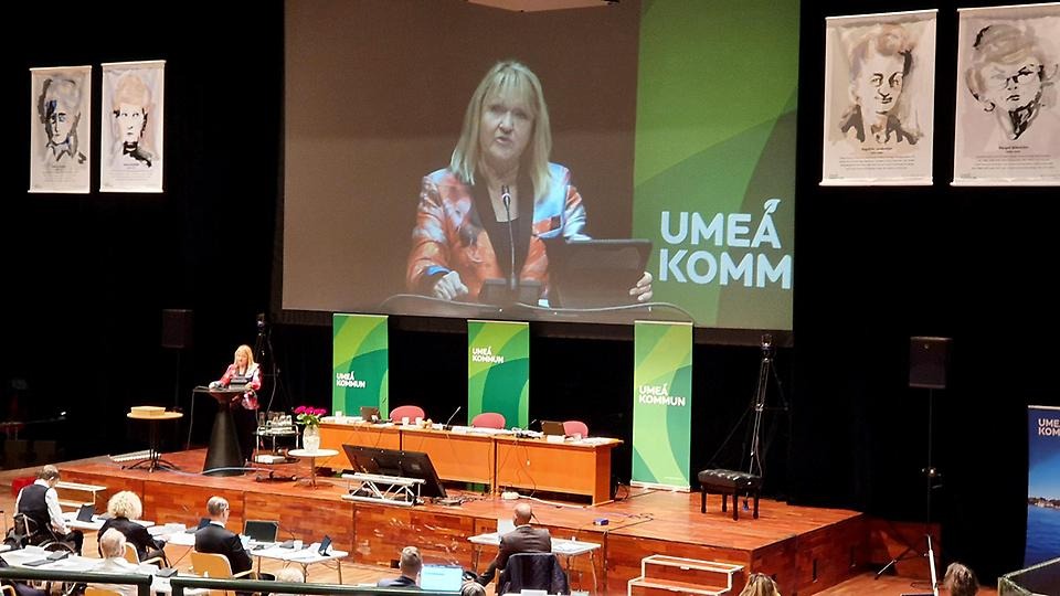 Marie-Louise Rönnmark talar under kommunfullmäktiges möte och uppmärksammar jämställdhet och demokrati, med vepor på kvinnliga förebilder i fonden.
