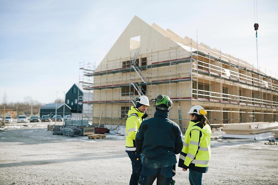 Tre byggnadsarbetare iklädda skyddskläder, varslarbeten och hjälmar står på en byggarbetsplats och pratar med varandra. I bakgrunden syns ett stort flervåningshus i trä under uppbyggnad, omgivet av byggnadsställningar.