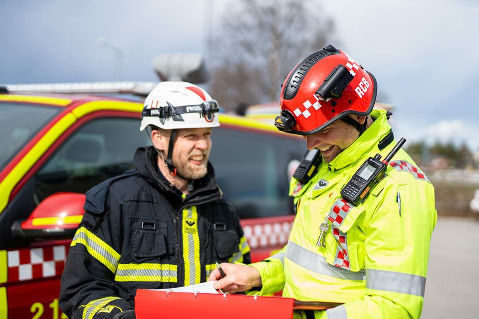 Två män i larmställ som står framför en brandbil, de tittar på varandra och skrattar