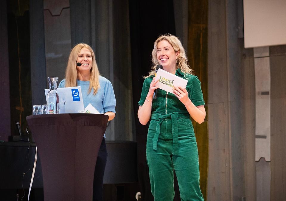 Två glada, unga kvinnor föreläser på en scen.
