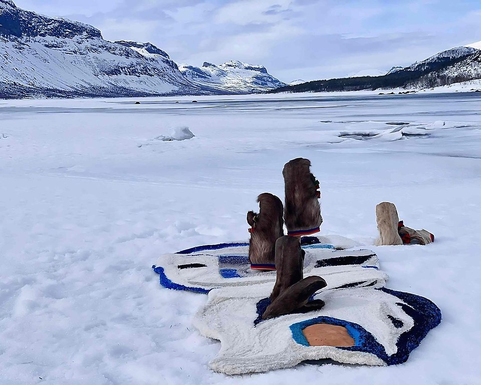Fjällvy med ett textilt konstverk i förgrunden. Konstverket består av traditionella samiska vantar i renskinn och en tuftad matta liknande en snödriva.