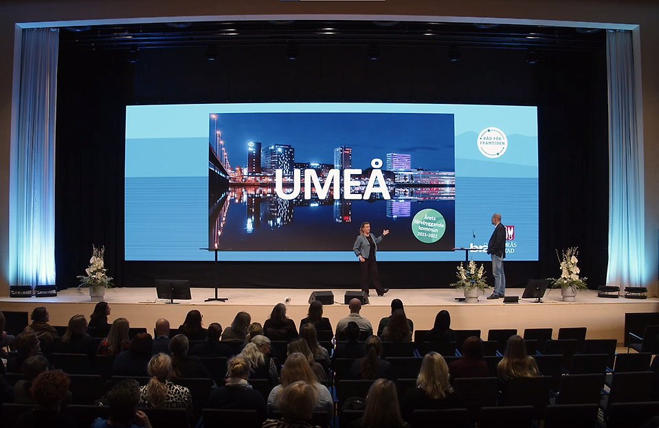 Två personer på en scen med stadsbild i bakgrunden med texten Umeå. I förgrunden syn publik i lokalen.