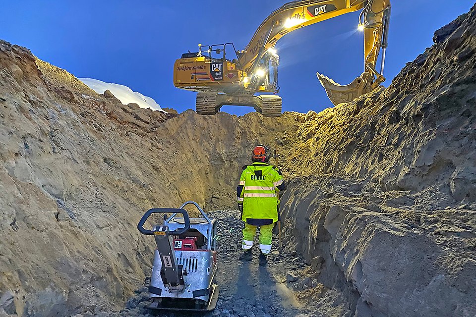 Bild nedifrån en grop, med en vägarbetare i förgrunden och en orange grävmaskin som står uppe vid gropens kant.