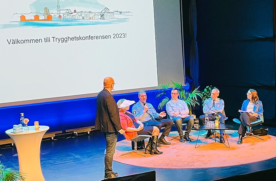 Fem personer sitter på en scen. En person står och det finns en skärm i bakgrunden med texten välkommen till trygghetskonferensen 2023.