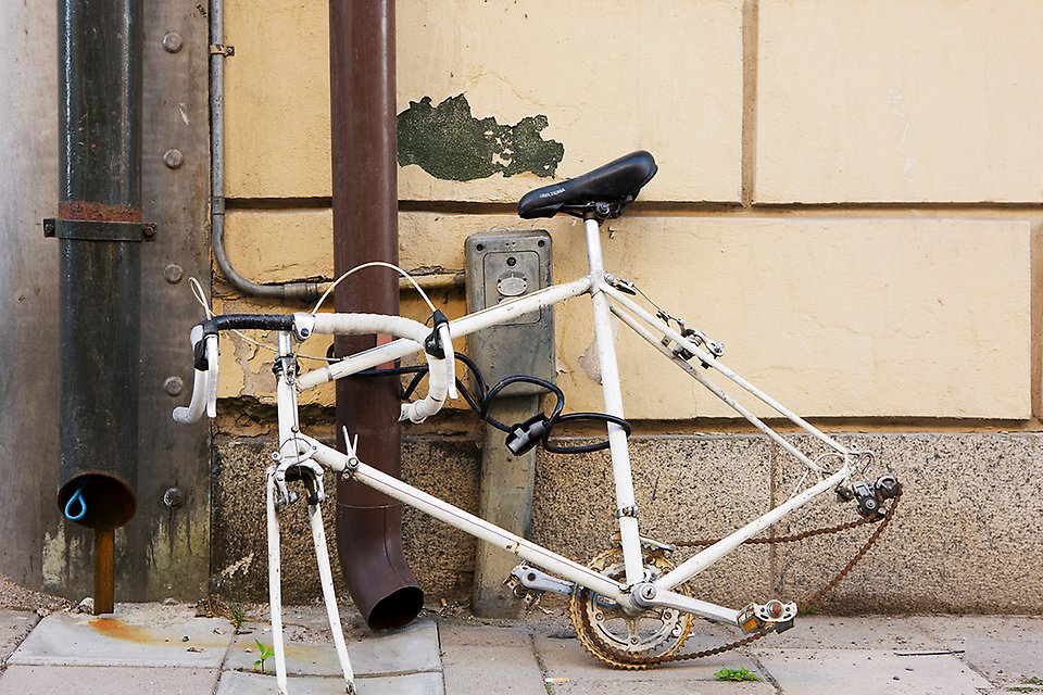 En trasig gammal cykel utan hjul står lutad mot en beige husvägg. Den är fastlåst i ett stuprör.