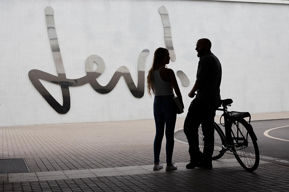 Siluetterna av en kvinna och en man med cykel som samtalar i gång- och cykeltunnel. Konstverket Lev! på tunnelväggen i bakgrunden.